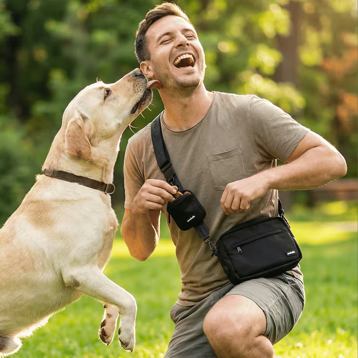 A cheerful man kneels on grass in a sunny Australian park, laughing as his yellow Labrador licks his face. He wears a casual tee and the Pawbella Crossbody Dog Walking Bag, with his pet backpack beside him.